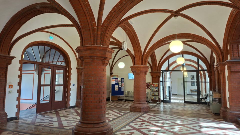 Innenansicht einer großen Halle mit roten Backsteinsäulen und kreuzrippenartigen Rundbögen, die eine helle, gewölbte Decke tragen. Der Raum hat ein gemustertes Fliesenbodenmosaik, hängende Kugelleuchten sowie Glastüren und Durchgänge zu angrenzenden Bereichen.