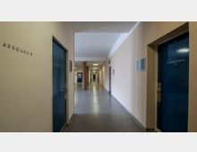 Ein langer, heller Flur in einem öffentlichen Gebäude mit beigen Wänden und blau lackierten Türen, darunter beschriftete Türen wie „Sitzungssaal 2“; die Beschilderung „Ausgang“ führt nach links. Der Boden ist mit grauem Linoleum ausgelegt, und im Hintergrund sind weitere Türen und ein Feuerlöscher erkennbar.