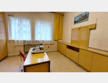 Ein schlicht eingerichtetes Büro aus der Vergangenheit mit Holzmöbeln: ein Schreibtisch mit Schreibmaschine, ein Stuhl, ein langer Wandschrank mit Schiebetüren und ein gerahmtes Bild an der Wand. Vor den Fenstern hängen weiße Gardinen und gelbe Vorhänge, darunter ist ein Heizkörper sichtbar.