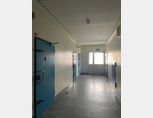 Langer, schmaler Flur in einem Haft- oder Sicherheitsgebäude mit mehreren massiven blauen Stahltüren auf beiden Seiten und glattem grauem Boden. Am Ende des Gangs befindet sich eine helle Doppeltür mit Fluchtweg-Schild darüber.