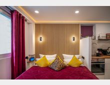 Modernes Schlafzimmer mit Akzentwand aus Holzlatten, beleuchtet durch zwei Wandleuchten. Ein Bett mit roter Decke und farbigen Kissen steht vor der Wand, angrenzend befindet sich eine kleine Küchenecke mit Elektrogeräten und Arbeitsfläche.