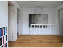 Flur eines modernen Hauses mit hellen Wänden, Holzboden und weißem Geländer. An der Wand hängt ein großes Waldmotivbild, daneben sind Türen mit Holzzargen sichtbar, ein Bücherregal steht links im Bild.