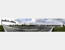 Panoramafoto eines Autobahnkreuzes mit mehreren Spuren und Abfahrten, Brücken und Verkehrsschildern. Im Hintergrund sind grüne Böschungen, Bäume und ein bewölkter Himmel sichtbar.