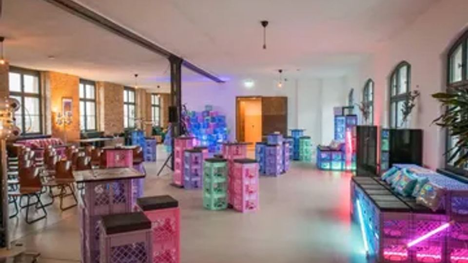 Großer, heller Innenraum mit hohen Decken, Stützsäulen und Bogenfenstern, eingerichtet mit Tischen, Stühlen und modularen Sitz- und Ablageelementen aus Kisten. Farbiges Neon- und LED-Licht beleuchtet einzelne Bereiche, mit Bar- bzw. Loungebereich entlang der Wand.