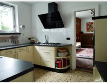 Moderne Küche mit cremefarbenen Unterschränken, schwarzen Arbeitsplatten und weiß gefliester Wand. Im Hintergrund ist ein angrenzendes Wohnzimmer mit rotem Teppich und Holzelementen zu sehen.