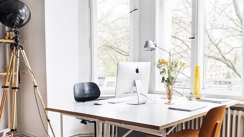 Bright office workspace with a large white desk on casters, two chairs, and a desktop computer, set on a parquet wood floor. Tall arched and rectangular windows fill the wall, with a vintage tripod floor lamp and a radiator beneath the windows.