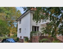 Zweistöckiges Wohnhaus mit weißer Fassade und rotem Ziegeldach, umgeben von Bäumen und Gartenbereich. Im Vordergrund ist eine Terrasse mit Geländer sowie ein geparktes Auto sichtbar.