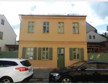 Zweistöckiges Wohnhaus mit orangefarbener Fassade, grünen Fensterläden und einer grünen Eingangstür, umgeben von weiteren Gebäuden; davor stehen zwei geparkte Autos auf einer Pflasterstraße.