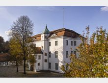 Historisches Gebäude mit weißer Fassade, roten Ziegeldächern und einem kleinen, grünen Kupferturm; umgeben von Bäumen mit herbstlich verfärbtem Laub auf einem gepflasterten Innenhof. Der Himmel ist klar mit vereinzelten Wolken.