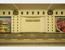 U-Bahn-Station mit der Aufschrift „Rüdesheimer Platz“ in einem weißen Wandmosaik. Die Wand ist von grünen Fliesen gesäumt und von seitlichen Wandmalereien mit Stadtszenen und Zügen flankiert.