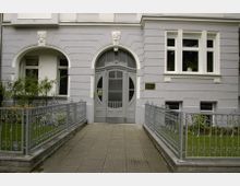 Fassade eines historischen Wohngebäudes mit grauem Putz, einem kunstvoll verzierten Rundbogenportal und einem schmiedeeisernen Gartentor. Vor dem Eingang befindet sich ein schmaler, gepflasterter Weg, links und rechts von kleinen Grünflächen und einem Metallzaun umgeben.