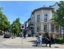 Historisches Eckgebäude mit heller Fassade und verzierten Fenstern, flankiert von einer gepflasterten Straße mit Gehwegen und vereinzelten Bäumen. Im Vordergrund sind Schilder für eine Halteverbotszone und einen Taxistand sowie einige Spaziergänger und geparkte Fahrzeuge zu sehen.
