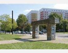Steinskulptur in Form eines Tors in einem Park mit gepflasterten Wegen und Grasflächen, im Hintergrund eine Straße mit parkenden und fahrenden Autos sowie mehrstöckige Wohngebäude mit bunten Fassaden.