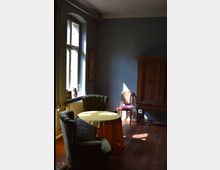 Wohnzimmer mit Holzboden und blau gestrichenen Wänden, eingerichtet mit zwei gepolsterten Sesseln, einem runden Tisch mit gelber Tischdecke, einem Holzkleiderschrank und einem Stuhl mit geflochtener Rückenlehne. Tageslicht fällt durch ein großes Fenster mit weißen Rahmen ein.