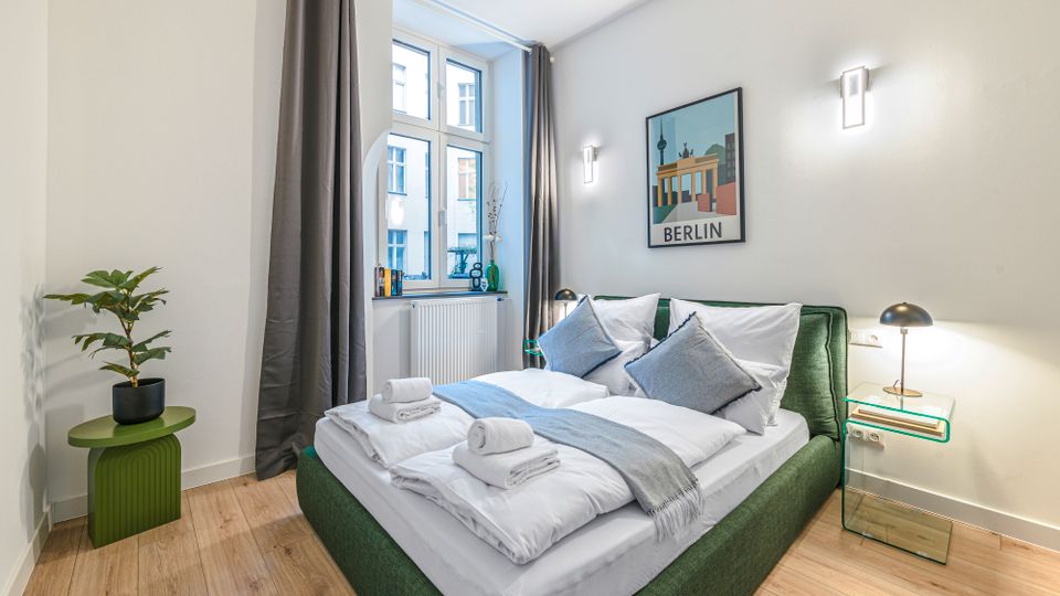 Helles, modern eingerichtetes Schlafzimmer mit grünem Polsterbett, weißer Bettwäsche und zwei Nachttischlampen. Große Fenster mit grauen Vorhängen, Holzfußboden sowie ein „Berlin“-Wandbild und ein kleiner Beistelltisch mit Pflanze ergänzen den Raum.