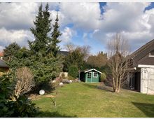 Garten mit grünem Rasen, mehreren Bäumen und Sträuchern sowie einer grünen Gartenhütte im Hintergrund. Rechts steht ein Haus mit dunklem Schrägdach und heller Fassade.
