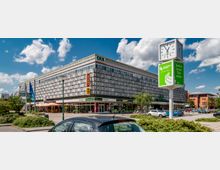 Einkaufszentrum mit markanter Fassadengestaltung aus rechteckigen Mustern. Im Vordergrund eine Uhr- und Werbesäule, parkende Autos und ein kleiner Grünbereich; Geschäfte und Restaurants im Erdgeschoss.