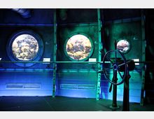 Ein Innenraum, der wie das Innere eines U-Boots gestaltet ist, mit metallischen Wänden und Bullaugenfenstern, die den Blick auf bunte Unterwasserwelten freigeben. Im Vordergrund befindet sich ein großes Steuerrad, das an die Steuerung eines Schiffs erinnert.