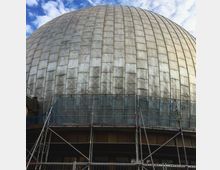 Silberfarbige Kuppel eines großen Gebäudes mit metallischen Platten, unterhalb von Baugerüsten und Netzen eingerahmt; der Himmel ist leicht bewölkt.