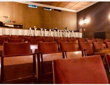 Innenraum eines historischen Kinos oder Theaters mit mehreren Reihen roter gepolsterter Klappsitze. Im Hintergrund ist eine erhöhte Bühne mit den weißen Buchstaben "ALHAMBRA" zu sehen, darüber eine hölzerne Decke und warme Beleuchtung.