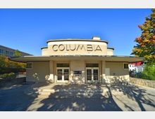 Eingang eines beige-farbenen Gebäudes mit dem Schriftzug „COLUMBIA” oberhalb der Türen. Das Design zeigt abgerundete Architekturformen mit einem breiten Vordach, Treppenstufen im Eingangsbereich und Bäumen im Hintergrund.