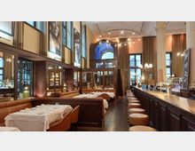 Innenraum eines eleganten Restaurants mit hoher Decke, großen Fenstern und Säulen. Die Einrichtung umfasst lederne Sitzbänke, Holztische mit weißen Tischdecken, eine lange Bar mit Barhockern und große Porträts an den Wänden.