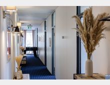 Ein schmaler, modern eingerichteter Flur mit blauen Teppichboden, weißen Wänden und dekorativen Trockenpflanzen in Vasen. Links sind Wandlampen und kleine Regale, rechts Türen und Fenster; am Ende des Flurs steht ein Kickertisch vor bodentiefen Fenstern mit Vorhängen.