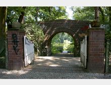 Backsteinbogenbrücke mit einem Weißmetalltor, flankiert von zwei gemauerten Pfeilern mit Kugeldekoration. Im Hintergrund befindet sich ein von Bäumen umgebener See.
