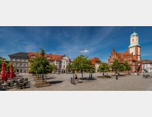 Großer gepflasterter Marktplatz mit Bänken, kleinen Bäumen, Laternen und einer Reihe von Wimpelgirlanden über dem Platz. Am Rand stehen mehrstöckige Wohn- und Geschäftshäuser sowie eine große Backsteinkirche mit hohem Turm und kupfergrünem Dach.