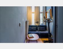 Schmaler Raum mit einer blauen Couch vor einer Wand mit mehreren hohen Fenstern, einem kleinen ovalen Couchtisch und einem Heimtrainer. Eine Wandleuchte und ein Heizkörper sind an der linken Seite sichtbar.