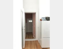 Schmaler Flur in einer Wohnung mit Holzdielenboden, einer Türöffnung mit weißen Türrahmen und einer offenen Tür links. Rechts im Vordergrund steht ein weißer Kühlschrank mit Geschirr auf der Oberseite, und die Wände des Flurs sind rosa gestrichen.