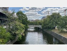 Fluss mit ruhiger Wasseroberfläche, umgeben von Bäumen und begrünten Ufern; im Hintergrund eine schmale Fußgängerbrücke, moderne Gebäude sowie Baukräne unter einem Himmel mit lockeren Wolken.