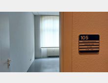 Eine offene Tür führt in einen leeren Raum mit grauem Boden und weiß gestrichenen Wänden. An der Wand neben der Tür hängt ein Schild mit der Aufschrift „105 Gesundheitsamt – Zahnärztlicher Dienst“ sowie mehreren Namen.
