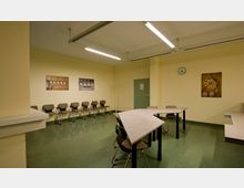 Schulungsraum mit gelben Wänden und grünem Bodenbelag. Stuhlreihen entlang der Wand, ein U-förmiger Tisch in der Mitte, eine Wanduhr und mehrere Bilder als Wanddekoration.