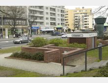 Außenansicht eines öffentlichen Damen-WCs mit Eingangsbereich aus roten Ziegelwänden, umgeben von niedrigen Sträuchern und Gehwegen. Im Hintergrund befinden sich eine mehrspurige Straße, geparkte und fahrende Autos sowie mehrstöckige Wohn- und Geschäftsgebäude.