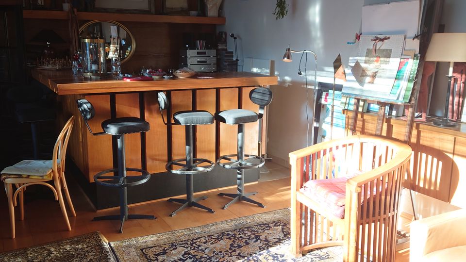 Innenraum eines gemütlichen Wohnraums mit einer hölzernen Bar, vier Barhockern und einem dekorativen Teppich auf einem Holzboden mit geometrischem Muster. Rechts befinden sich ein Holzstuhl mit rotem Kissen, ein Bücherstapel und Staffeleien, während links ein zweiter Teppich liegt.