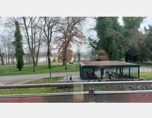 Ein Park mit einer überdachten Fahrradabstellanlage im Vordergrund, umgeben von gepflasterten Wegen, Rasenflächen und kahlen Bäumen. Im Hintergrund sind ein Fluss und eine Baumreihe sichtbar.