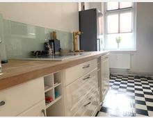 Helle Küche mit weißen Schränken, einer Holzarbeitsplatte, Glasrückwand und einem schwarz-weiß karierten Boden. Im Hintergrund sind ein großes Fenster mit Jalousien, ein grauer Kühlschrank und Heizkörper zu sehen.