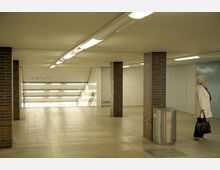 Unterirdischer U-Bahn-Zugang mit gefliesten Wänden in Weiß und Mosaik-Türkis, beleuchtet durch helle Deckenlichter. Im Vordergrund stehen gemauerte Säulen und ein silberner Abfalleimer, eine ältere Frau mit heller Jacke läuft rechts vorbei.