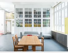 Ein heller Innenraum mit großen weißen Sprossenfenstern, einer grauen Couch und einem Holztisch mit Stühlen im Vordergrund. Die Rückwand ist mit Blumenmustern dekoriert und durch Glaselemente unterteilt, während seitlich hohe Fenster viel Tageslicht einlassen.