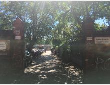 Eingang zu einem gepflasterten Innenhof, flankiert von zwei Backsteinpfeilern mit befestigten Schildern, die Adressen und Geschäftsnamen anzeigen. Der Hof ist von Bäumen beschattet, im Hintergrund sind geparkte Fahrzeuge und Gebäude zu sehen.