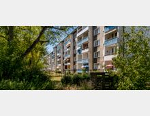 Mehrstöckige Wohngebäude mit Balkonen, die mit Sonnenschirmen und Blumenkästen dekoriert sind, umgeben von Bäumen und dichter Vegetation. Im Vordergrund ist ein Naturbereich mit Gras und Sträuchern zu sehen.