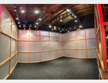 Ein Indoor-Raum mit holzgerahmten, lichtdurchlässigen Kunststoffwänden und einem dunklen Teppichboden. Die Decke besteht aus sichtbaren Holzbalken und ist mit mehreren modernen Strahlern ausgestattet.