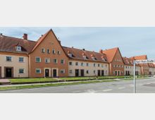 Reihenhauszeile mit orangefarbenen und beigefarbenen Fassaden, roten Satteldächern und kleinen Vorgärten. Im Vordergrund verläuft eine Straße, an der ein Straßenschild mit der Aufschrift "Karl-Thon-Straße" steht.