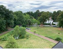 Blick auf eine grüne Parklandschaft mit Rasenflächen, Bäumen und einem kleinen Teich in der Mitte. Im Hintergrund sind eine Straße, mehrere parkende Autos und ein weißes Wohnhaus zu sehen, umgeben von weiteren Bäumen.