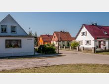 Eine ruhige Wohnsiedlung mit mehreren Einfamilienhäusern, die rote Ziegeldächer und weiße oder farbig gestrichene Fassaden besitzen. Der gepflegte Bereich zeigt gepflasterte Wege, Vorgärten mit Pflanzen und klare Wegweiser unter blauem Himmel.