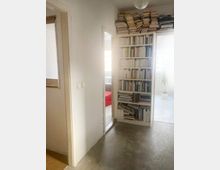 Ein schlichter Flur mit weiß gestrichenen Wänden und Betonboden. Rechts steht ein hoher Bücherregal, überladen mit Büchern, einige Bücher sind auch oben gestapelt. Im Hintergrund sind zwei Räume sichtbar, einer mit einem roten Sofa und der andere mit einer Pflanze am Fenster.