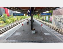 Bahnhof mit freistehendem Mittelbahnsteig, überdacht durch eine Holzkonstruktion. Zwei Gleise führen links und rechts am Bahnsteig entlang, während digitale Anzeigetafeln, Werbeplakate an einer Backsteinmauer und grüne Vegetation im Hintergrund sichtbar sind.