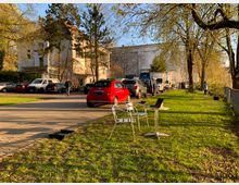 Eine Grünfläche mit Bäumen und einem kleinen Tisch mit Metallstühlen im Vordergrund. Im Hintergrund stehen Autos und Transporter auf einer gepflasterten Einfahrt neben einem hellen Gebäude mit roten Dachziegeln.