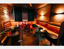 Innenraum einer Lounge oder Bar mit freiliegenden Backsteinwänden, gepolsterter Leder-Sitzbank entlang der Wände und mehreren kleinen runden Tischen mit Stühlen. Gedämpfte Wandlampen und dunkle Vorhänge sorgen für eine warme Beleuchtung, daneben führt ein Treppenabgang mit Geländer nach unten.